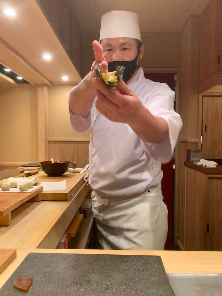Chef at Sushi Yuu extending a hand-roll across the counter with finger pointed